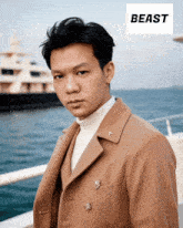 a man wearing a tan coat and a white turtleneck stands in front of a boat with the word beast above him