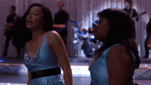 two women in blue dresses stand on a stage with a band in the background
