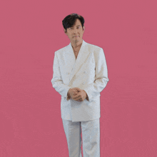 a man in a white suit stands with his hands folded in front of a pink background