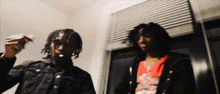 two men are standing next to each other in a room . one of the men is smoking a cigarette .