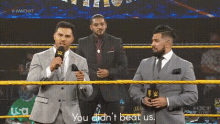a man in a suit is talking into a microphone in a wrestling ring while another man stands behind him .