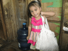 a little girl in a pink dress holds a white bag in front of a bottle of water