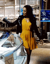 a woman in a yellow dress and black jacket is standing in a room with her arms outstretched .