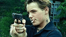 a young man with a tattoo on his neck is smoking a cigarette and pointing a gun at the camera .