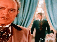 a man in a suit and tie is standing in front of a blue curtain with the words picplaypost written on the bottom