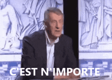 a man in a suit says " c'est n'importe " in front of a statue