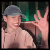 a woman wearing a baseball cap and a hoodie is laughing and waving at someone .