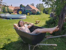a naked man is laying in a wheelbarrow with a tractor in the background