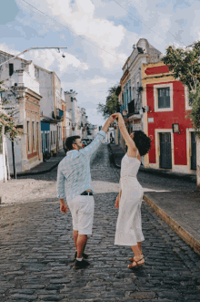 a man and woman are dancing on a cobblestone street with a red building in the background