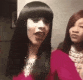 two women are standing next to each other in a room . one of the women is wearing a wig .