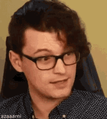 a man wearing glasses and a polka dot shirt is sitting in a chair and making a funny face .