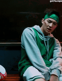 a man wearing a green bandana and a green hoodie sits on a bench next to a soccer ball