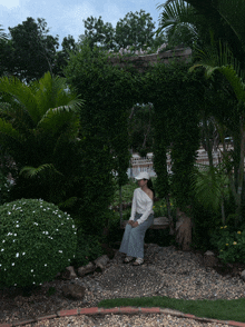 a woman in a white hat sits on a bench under a lush green archway