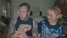 a man with a tattoo on his forehead is standing next to a woman wearing a gifrun.com shirt