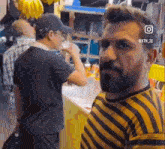 a man with a beard is standing in front of a counter while another man drinks from a bottle .