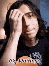 a man with long hair is holding his hand to his forehead and says ok waiting