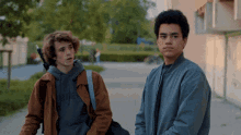 a boy wearing a hoodie that says rue stands next to another boy wearing a bomber jacket