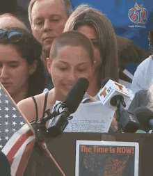 a woman stands in front of a microphone with a sign that says the time is now