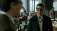 a man in a suit and glasses is smiling in an office while talking to another man .