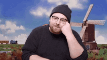 a man wearing glasses and a beanie looks at the camera with a windmill in the background