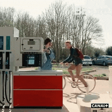 a man riding a bicycle at a gas station with canal + written on the bottom