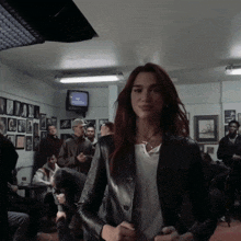 a woman in a leather jacket stands in a crowded room with a tv on the wall