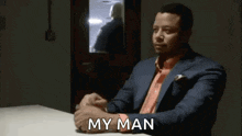 a man in a suit is sitting at a table with his hands folded and the words `` my man '' written on the table .