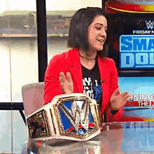 a woman in a red jacket is sitting at a table with a wrestling championship belt on it .