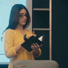 a woman in a yellow shirt and glasses is holding a notebook in her hands .