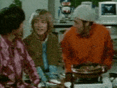 a group of people are sitting around a table with a man in an orange jacket cooking