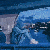 a person in a blue hoodie sits on a bridge overlooking a body of water