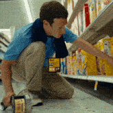 a man in a blue shirt is kneeling down in a grocery store aisle looking at cereal