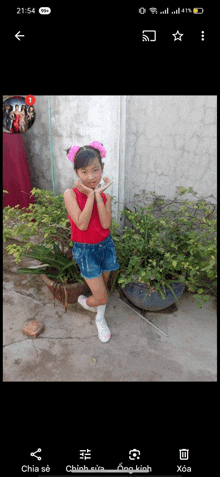 a little girl wearing a red top and blue shorts stands in front of a plant