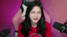 a woman in a red shirt is sitting in a pink chair next to a microphone and making a funny face .