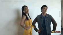 a man in a denim jacket stands next to a woman in a yellow dress with a flower in her hair ..