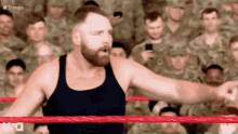 a man with a beard is standing in a wrestling ring with soldiers in the background .
