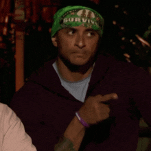 a man wearing a green survivor headband points at something