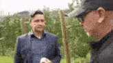 two men are standing in a field talking to each other . one of the men is wearing a hat .