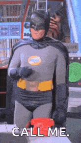 a man in a batman costume is standing in front of a red telephone and saying `` call me '' .
