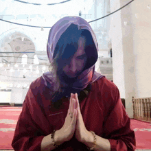 a woman wearing a red shirt and a purple scarf prays in a mosque