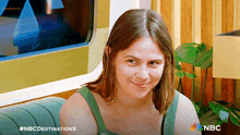 a woman in a green tank top is smiling in front of a sign that says nbc destination x