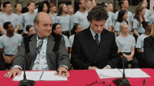 two men are sitting at a table with microphones in front of a crowd of people wearing white shirts with the letters ncs