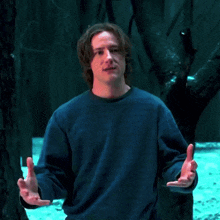 a man in a blue sweater is standing in front of trees with his hands folded