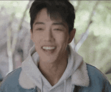 a young man wearing a hoodie and a denim jacket smiles with his eyes closed