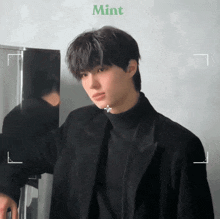 a young man wearing a black jacket and turtleneck is standing in front of a fridge .
