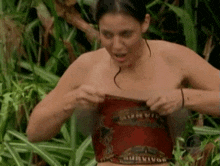 a woman is standing in the jungle without a shirt on and taking off her top .