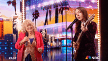 a woman singing into a microphone next to an older woman with a guitar