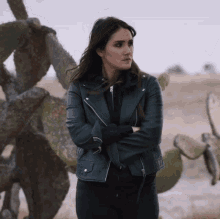a woman wearing a black leather jacket stands in front of a cactus