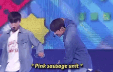 two men are dancing on a stage with the words pink sausage unit written above them .