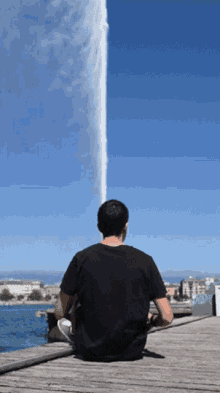 a man in a black shirt sits on a dock looking at a fountain